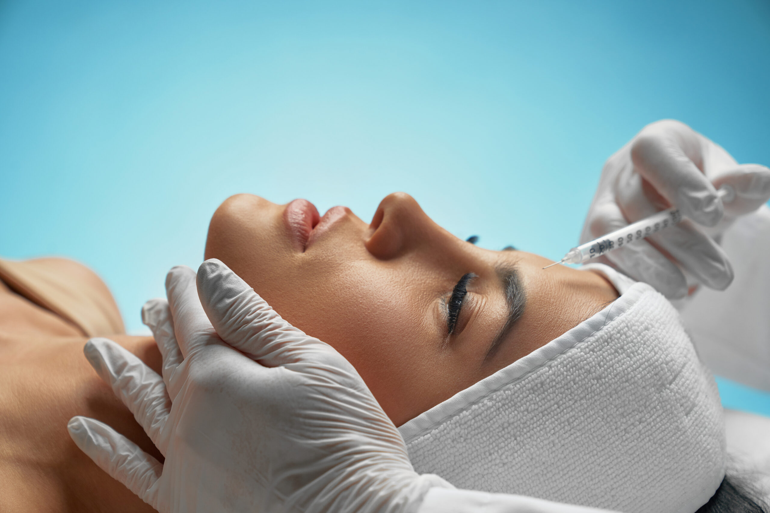 Cosmetic botox injection in female forehead. Close up of botox injection in female forehead. Side view of cosmetologist using syringe with special liquid, holding chin while patient with towel on head lying. Concept of cosmetology, beauty.